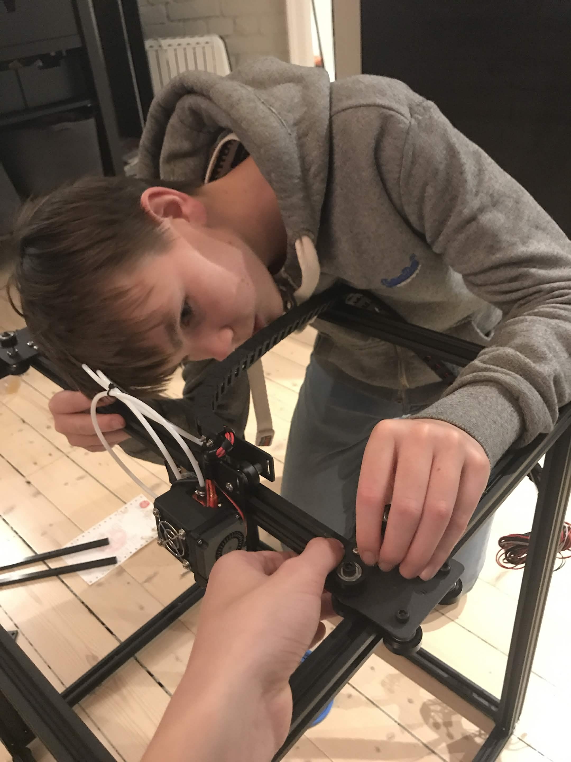 A teenager hunched over an aluminium-profile printer frame, tightening a belt on the X-axis carriage. Filament extruder visible beside him.