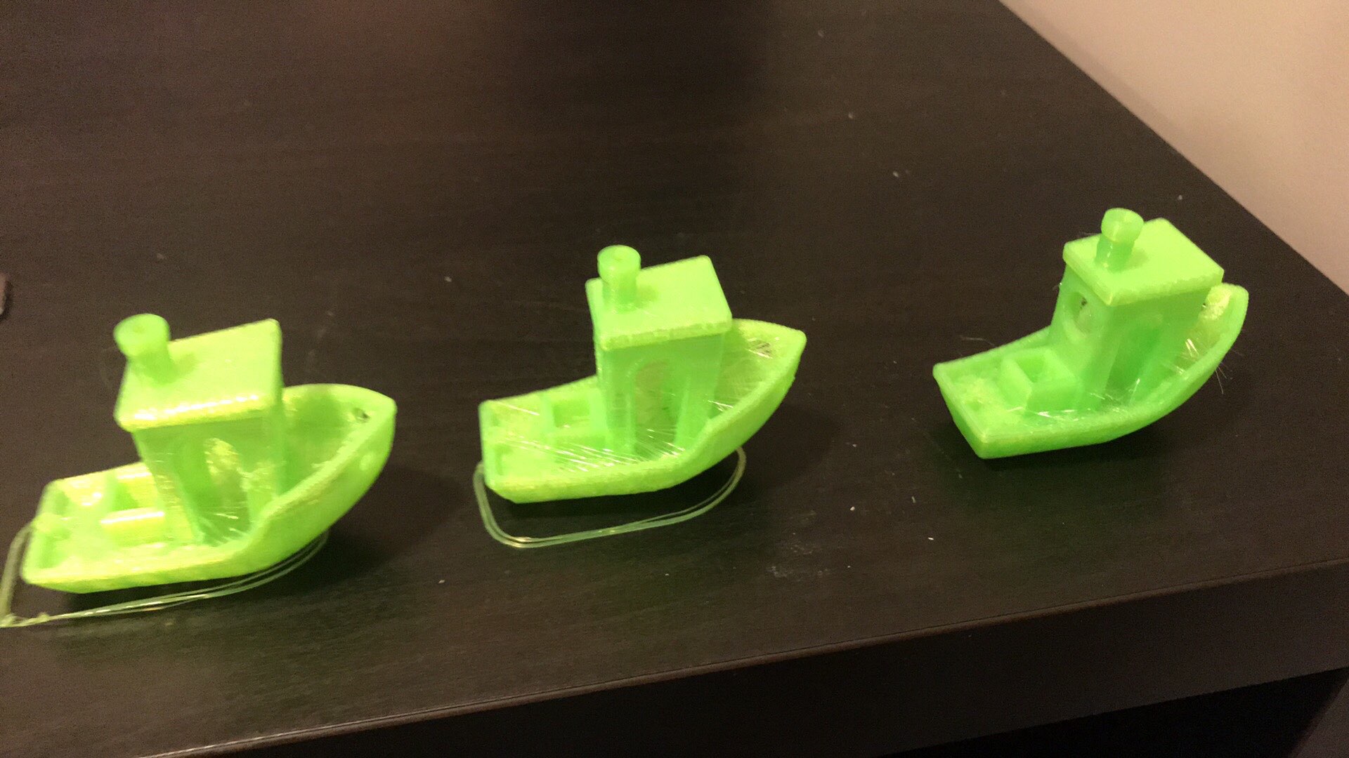Three green benchy boats lined up on a dark desk. Each one shows the same model printed with slightly different settings, with different amounts of stringing and surface roughness on the hull and roof.