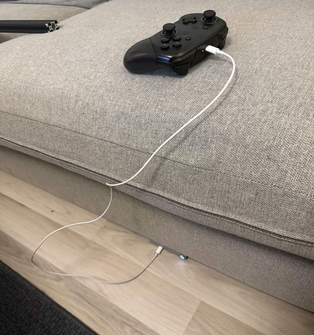 A Nintendo Switch Pro controller resting on a couch cushion, charging via a white cable that runs down to the dock built into the couch below.
